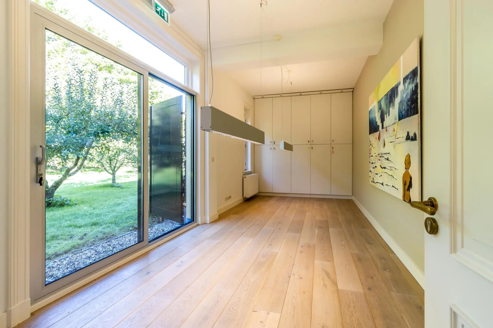 Lichte kamer met houten vloer, kunst aan de muur, ingebouwde kasten en uitzicht op een groene tuin via glazen schuifdeuren.